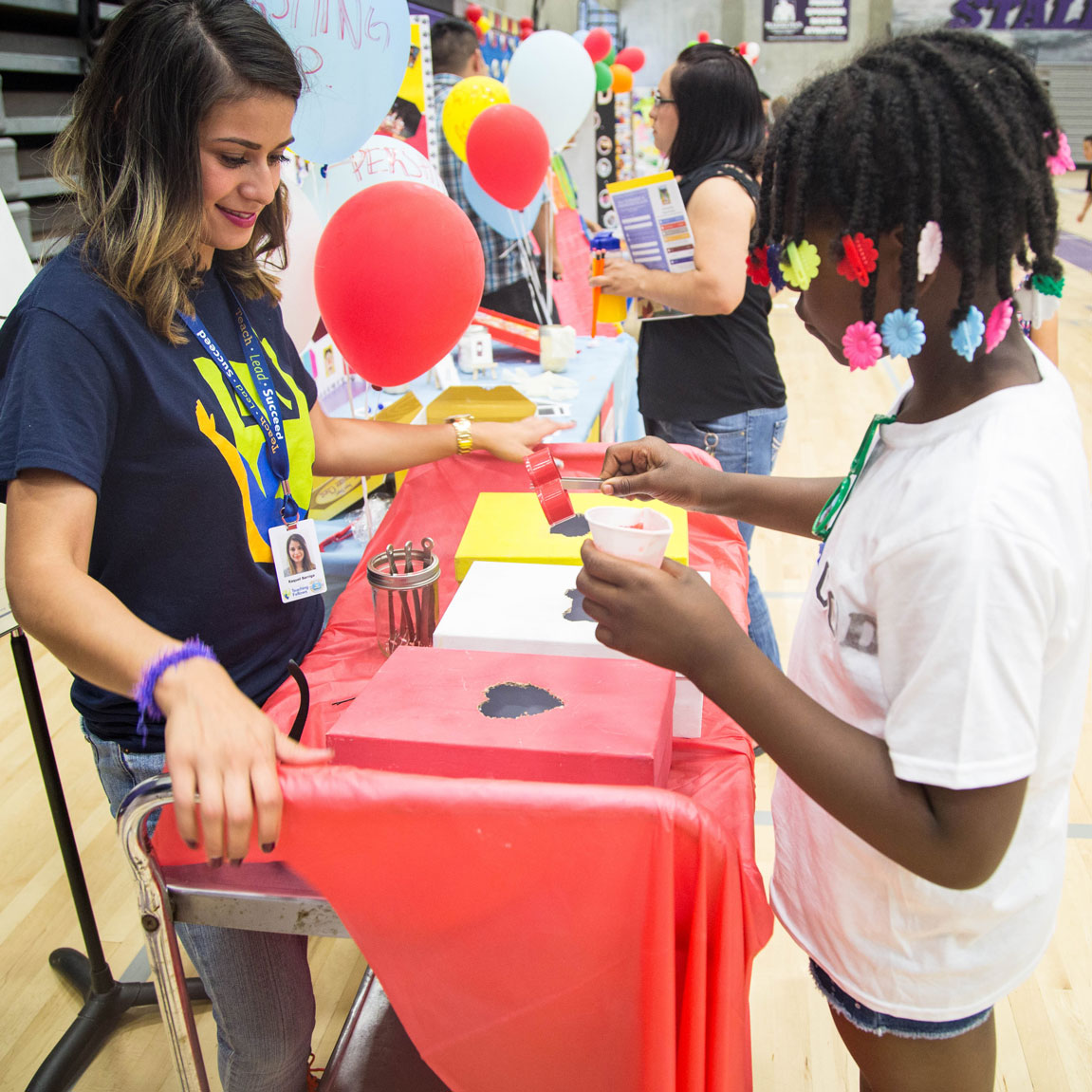 Closeup of a Teaching Fellow standing at an activity booth with a red tablecloth and a student with black hair and multiple colorful hair clips participating in the activity.