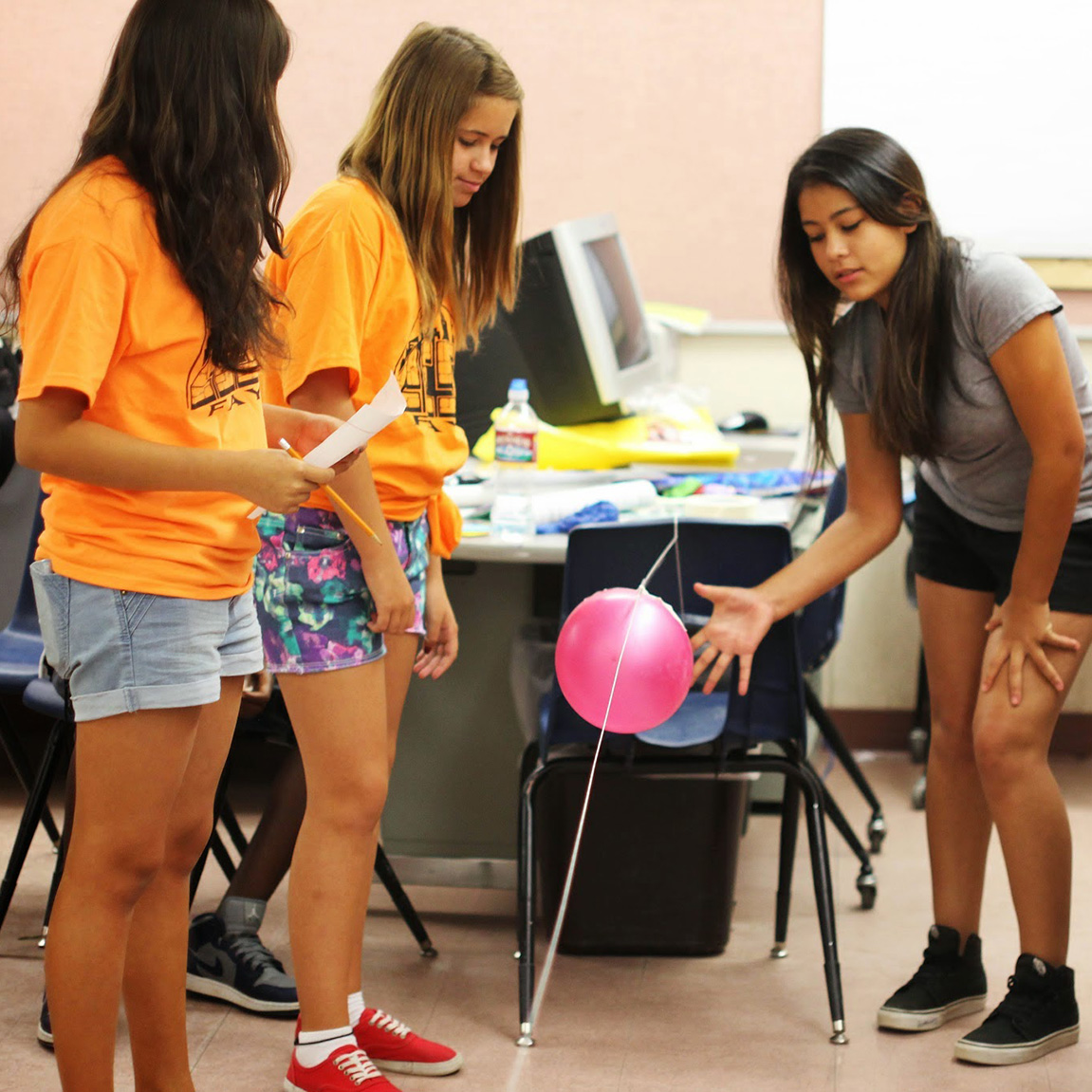Three students, with two, wearing matching orange t-shirts and one wearing a gray t-shirt, participating in an activity with a pink balloon moving along a string.