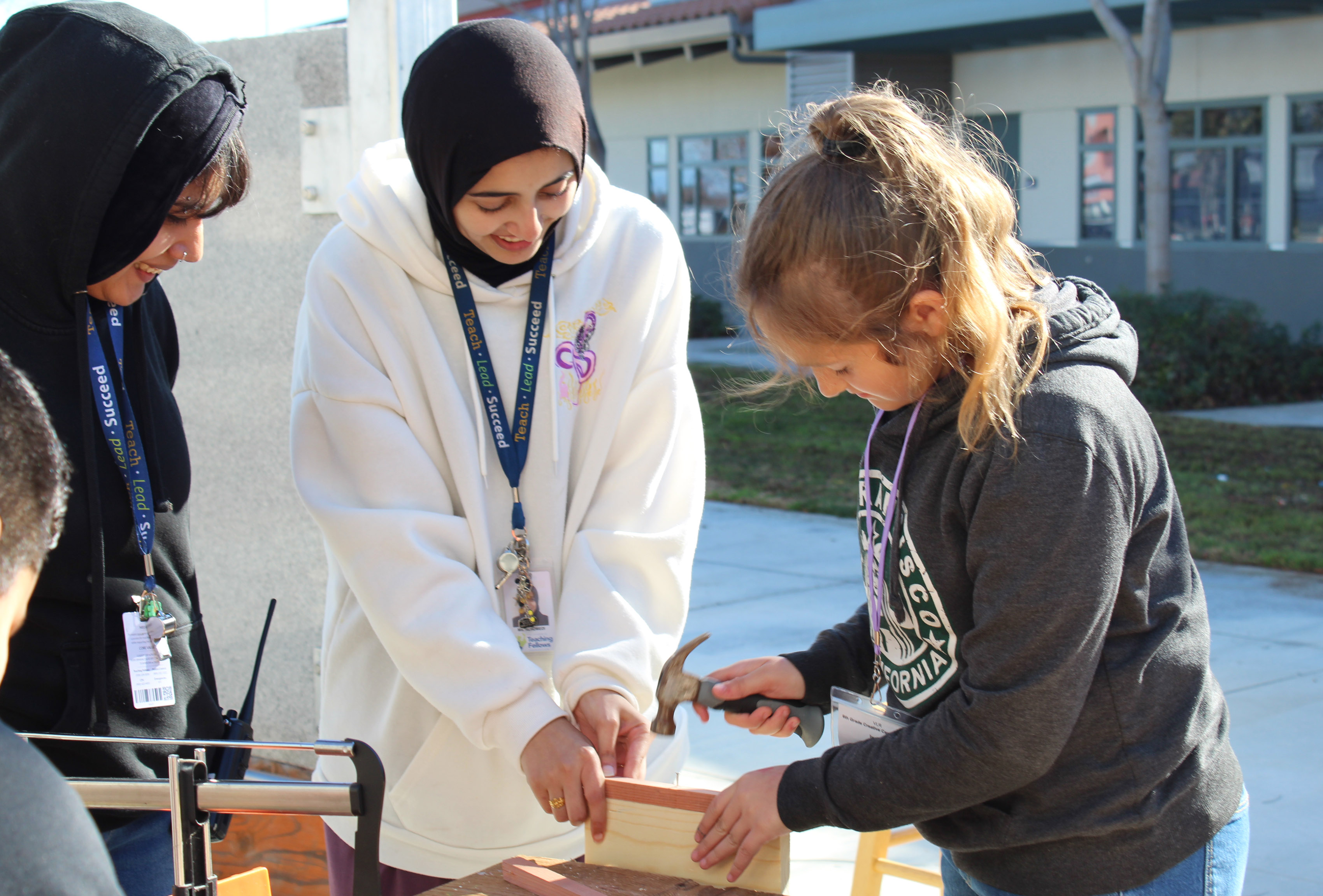Woman wearing a white hoodie working with student using a hammer.
