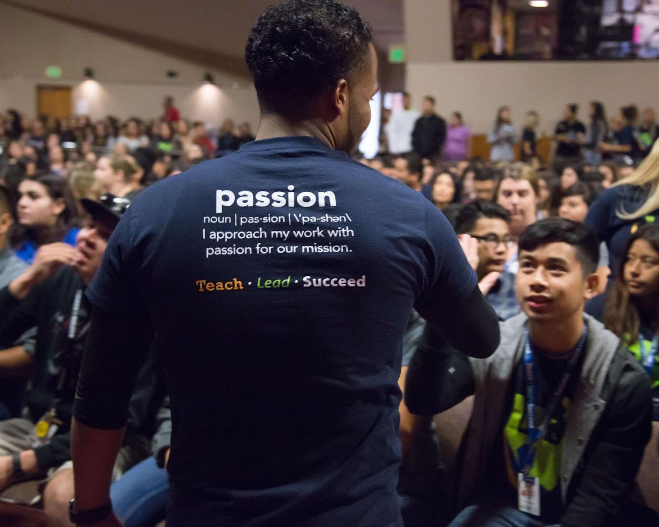 Image of a male with back against screen, greeting another male. The back of his shirt has the word Passion.