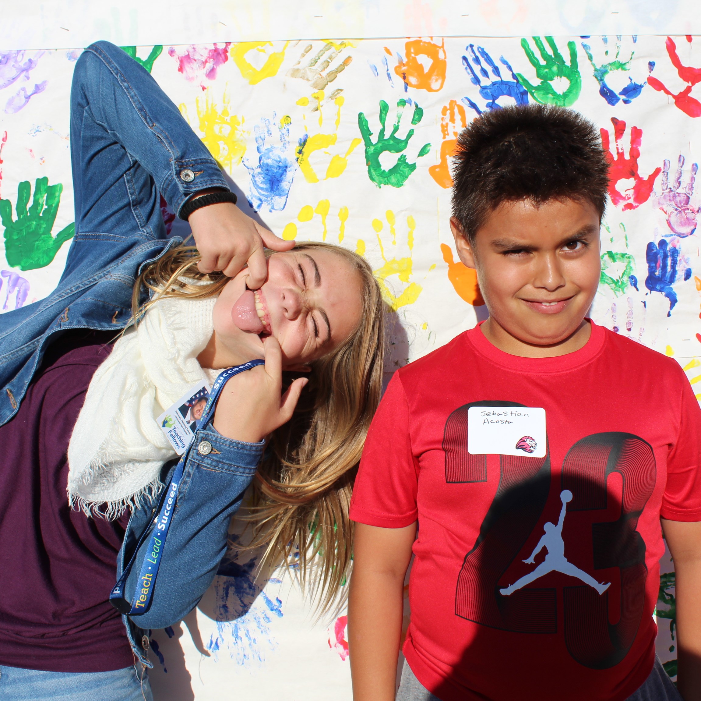 A Teaching Fellow poses with a silly face alongside a young male student in a red shirt in front of a backdrop full of painted hands in a rainbow of colors.