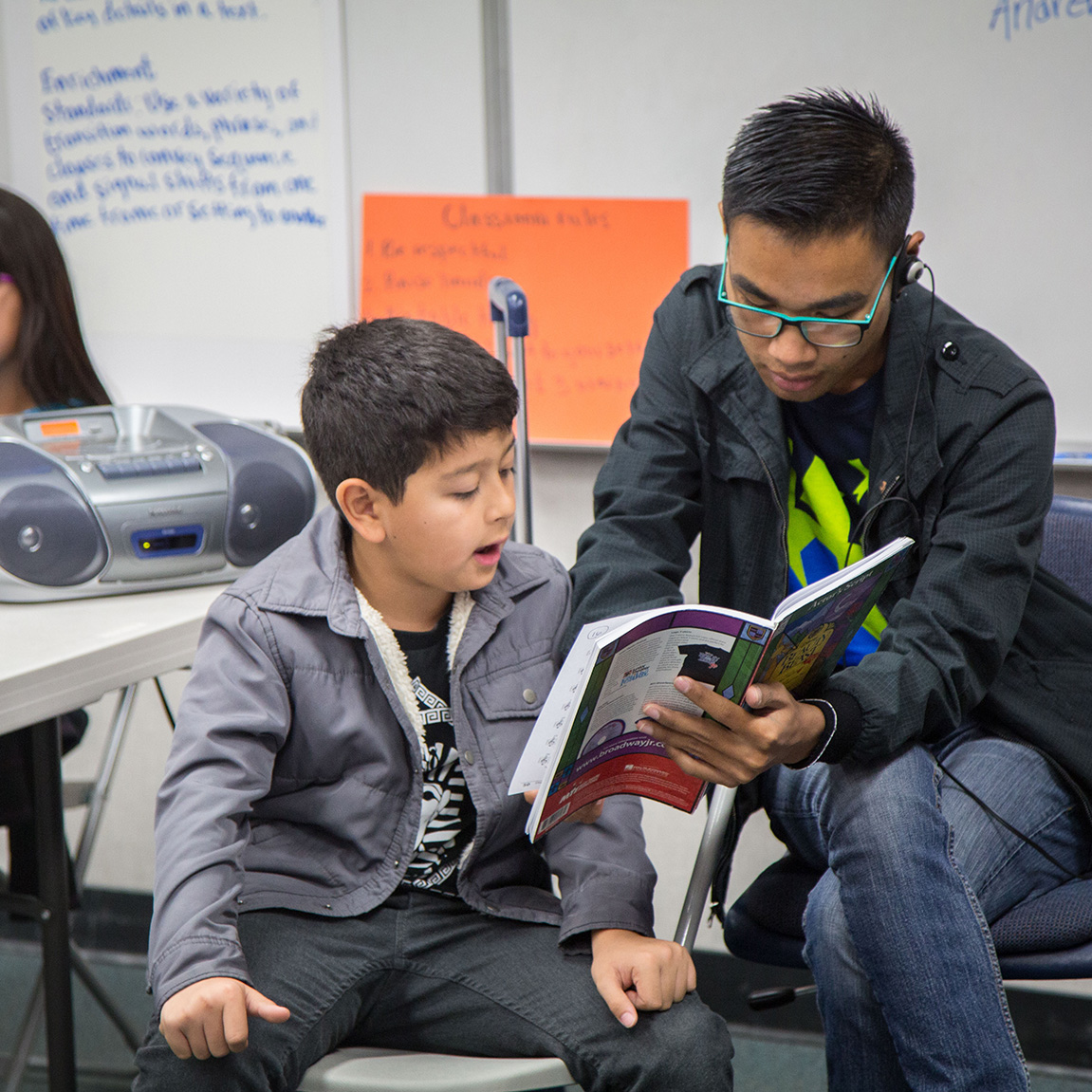 A closeup of a male Teaching Fellow tutor in a CTFF t-shirt and black jacket holding a book while a young male student follows along and reads out loud.