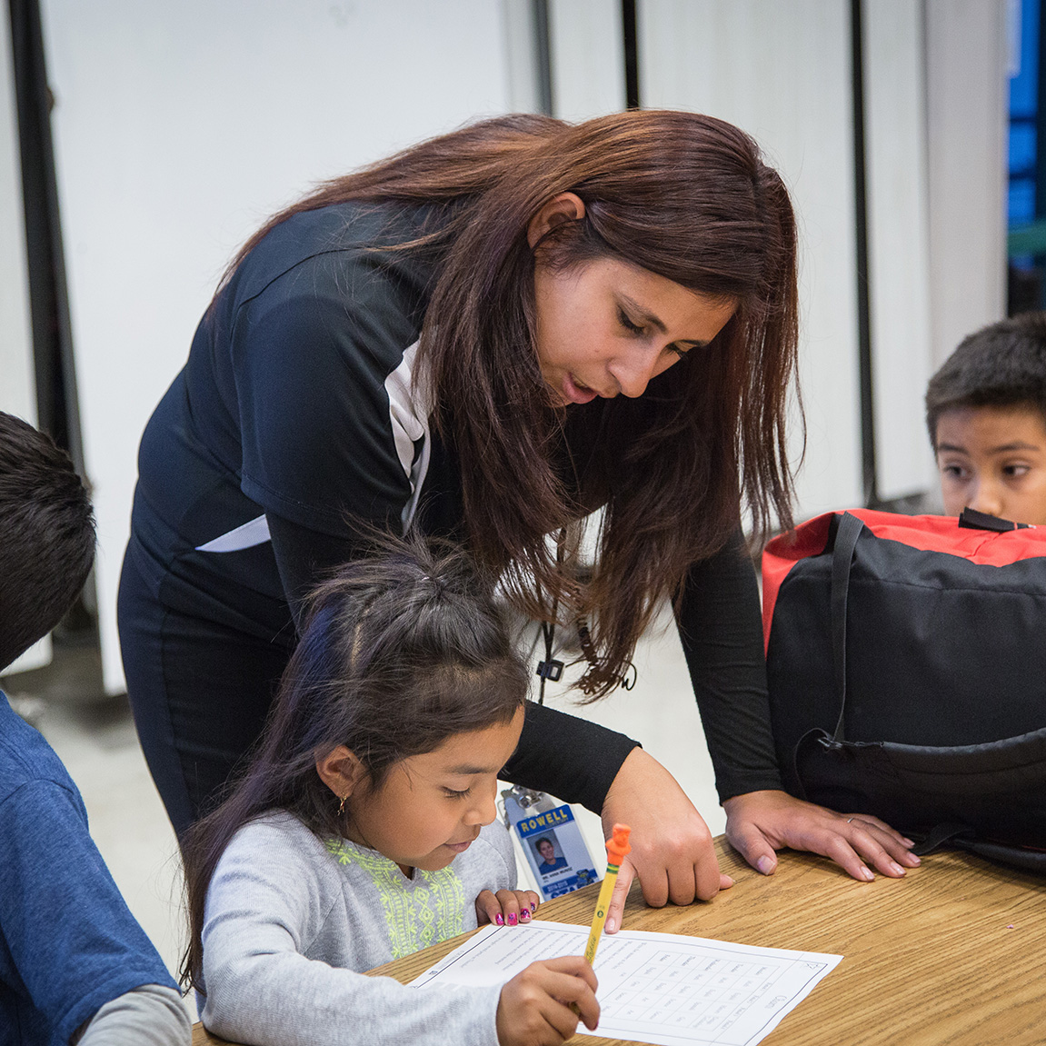 A closeup of a Teaching Fellow tutor leaning down and helping a student with a worksheet with another student looking on from a distance.