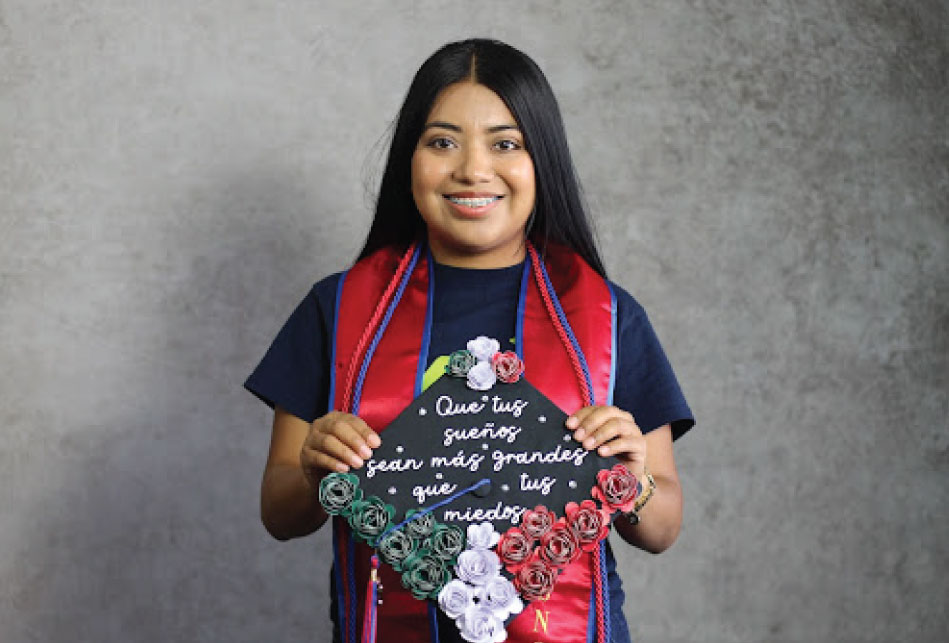 College student standing in front of grey wall wearing a Teaching Fellows tshirt and red graduation stole with red and blue ropes, holding a decorated graduation cap with a Spanish phrase written on it and green, white, and red roses attached to the border.