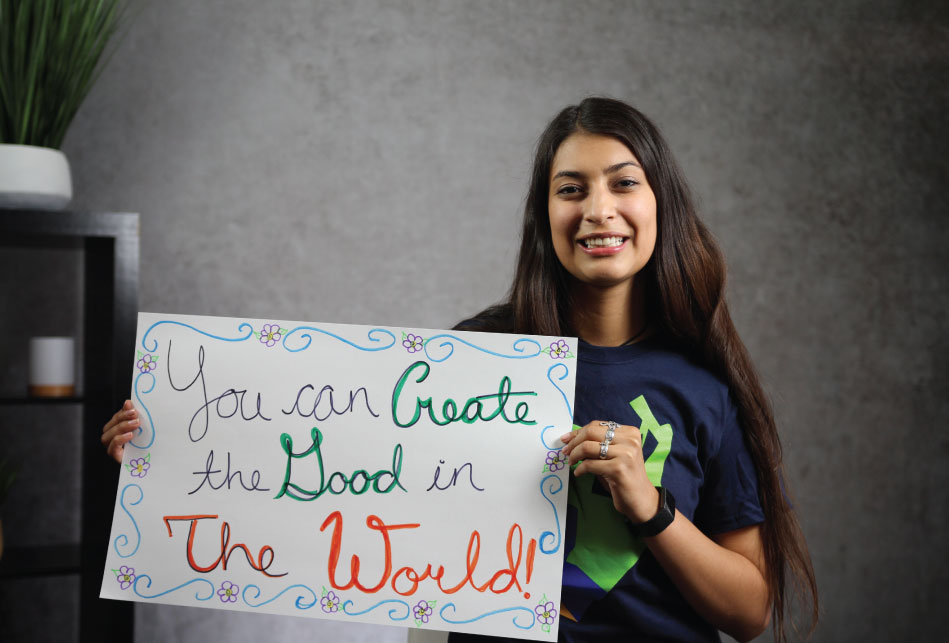Woman with long dark hair smiling and holding a handmade sign that says YOU CAN CREATE THE GOOD IN THE WORLD with flowers and swirls as the border images.