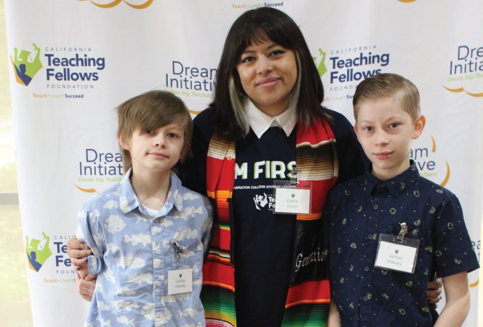 Woman wearing a shirt that says I’M FIRST standing in front of a white photo backdrop with her arms around her two sons on either side of her, both wearing collared shirts.